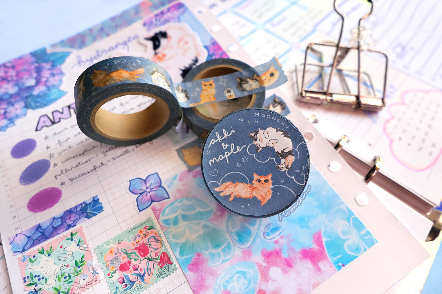original artist washi tape by moonchu as a tribute to two of her cats, akki and maple. this photo showcases the packaging label with one illustration of akki and one illustration of maple on a navy/blue background with white cloud and star doodles. the rest of the flatlay consists of other cute stationery items and journal pages.