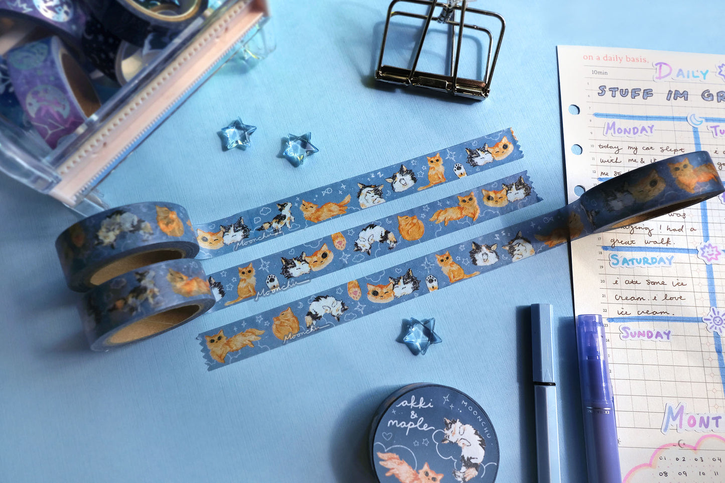 original artist washi tape by moonchu as a tribute to two of her cats, akki and maple. three washi tape are unrolled for the viewer to see the various kitty designs (both of their faces smushed together and smiling, a calico paw and an orange kitty paw, akki and maple in sleeping/sitting/yawning poses). orange cat and calico cat are drawn in various poses on a navy/blue background with white doodles (such as clouds and stars and hearts). the rest of the flatlay consists of other cute stationery items.