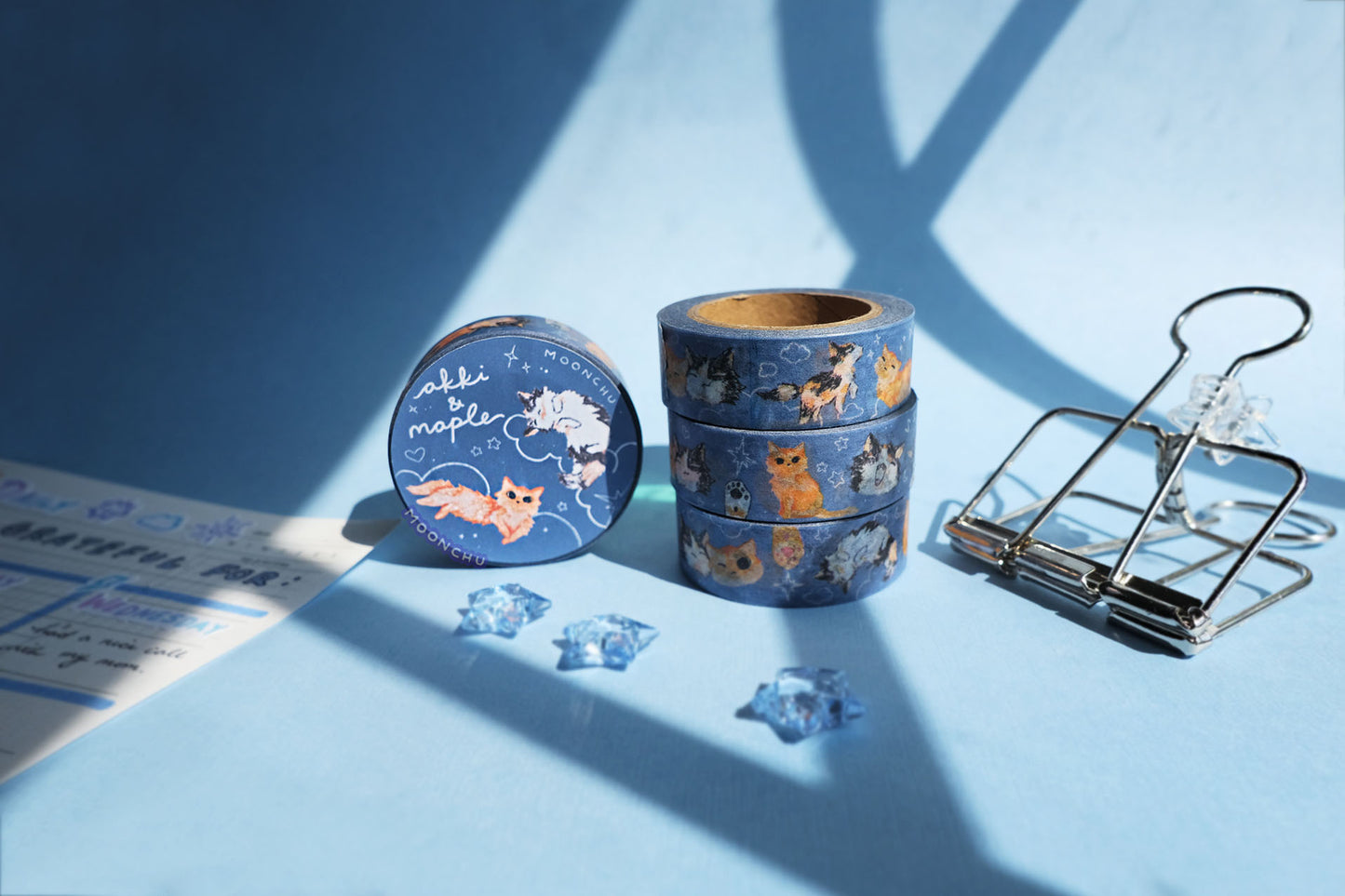 original artist washi tape by moonchu as a tribute to two of her cats, akki and maple. three washi tape are stacked atop one another, and one is on the side showcasing the packaging label. orange cat and calico cat are drawn in various poses on a navy/blue background with white doodles (such as clouds and stars and hearts). the backdrop is a pretty blue with sunlight streaming through.