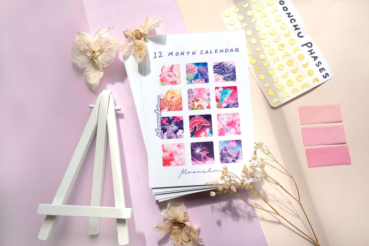 flatlay image with thumbnail previews of moonchu's original mystical calendar design. each month depicts moonchu's interpretation of that month's full moon meaning. vibrant, saturated, surreal and nature inspired watercolour and mixed media paintings recreated in high quality prints via monthly calendar. also shows gold moon phase stickers to embellish your calendar with to depict the changing moons.