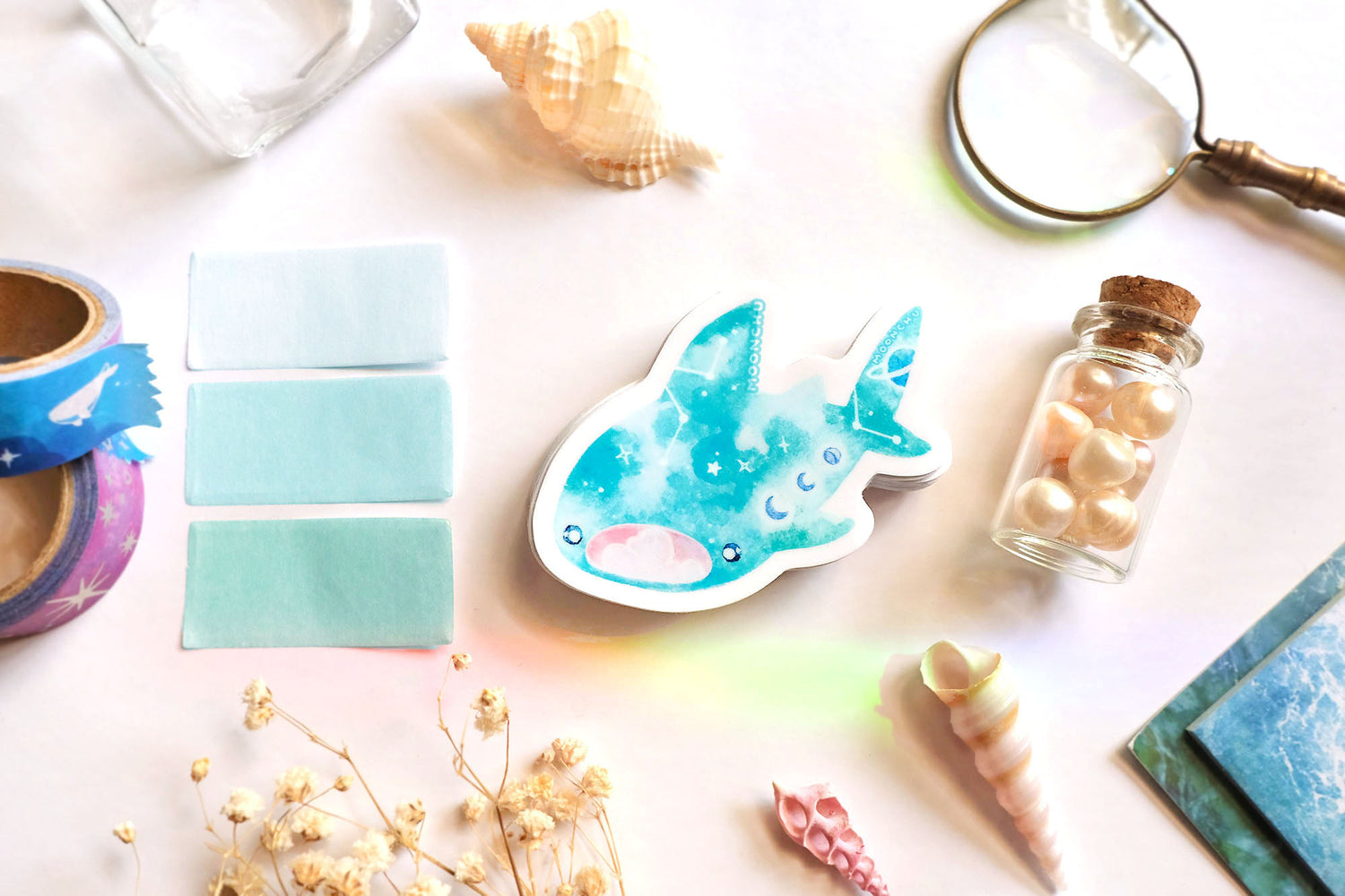 original art illustrated sticker by moonchu depicting a watercolour painted whale shark! mouth ajar, with colours of cyan and blue, it has little constellations and moon phases upon its body. it also has saturn on its tail! flatlay consists of other ocean items, like little shells and pearls. a high quality vinyl sticker for shark lovers or ocean gifts. perfect sticker for shark week.