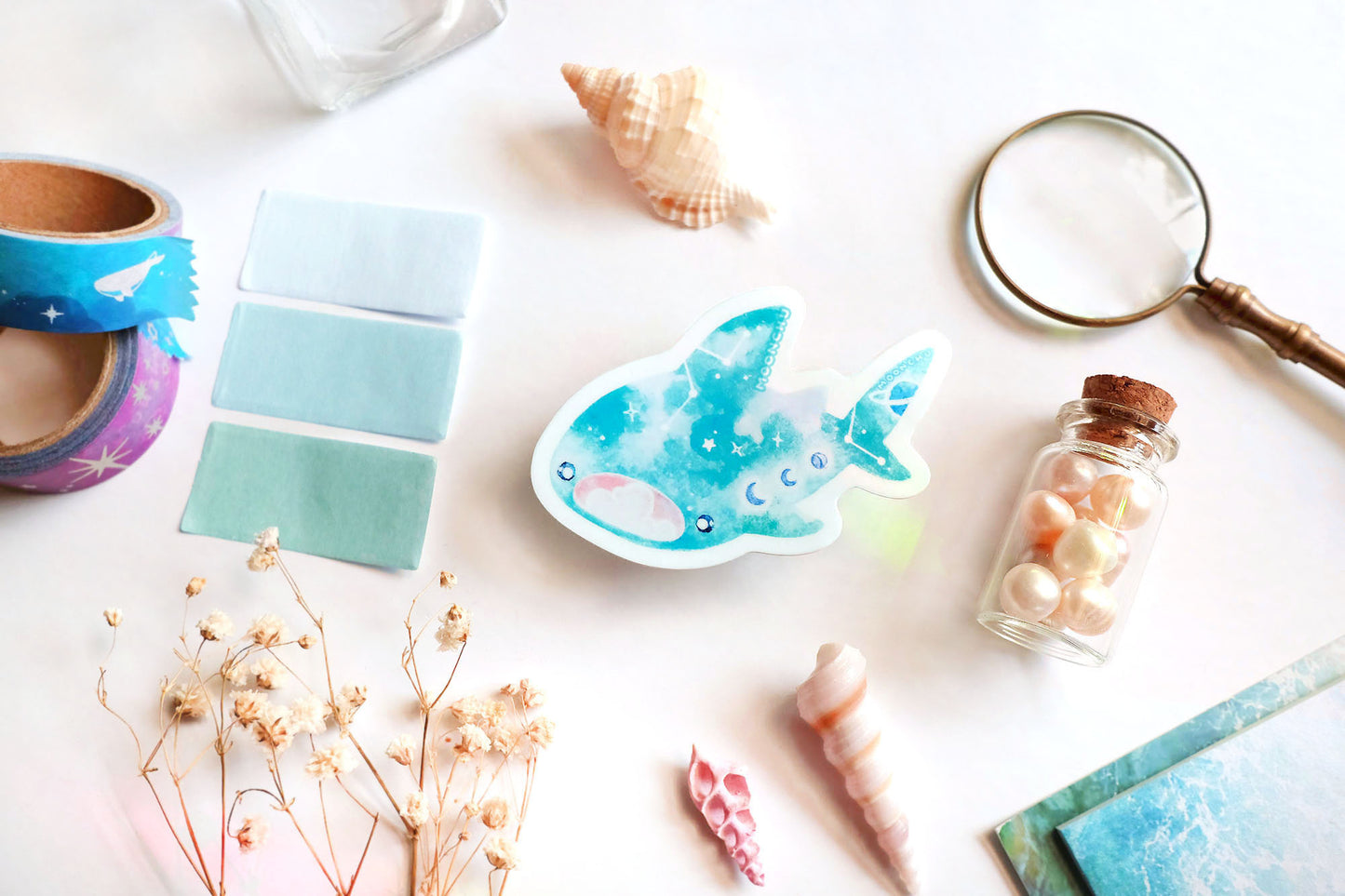 original art illustrated sticker by moonchu depicting a watercolour painted whale shark! mouth ajar, with colours of cyan and blue, it has little constellations and moon phases upon its body. it also has saturn on its tail! flatlay consists of other ocean items, like little shells and pearls. a high quality vinyl sticker for shark lovers or ocean gifts. perfect sticker for shark week.