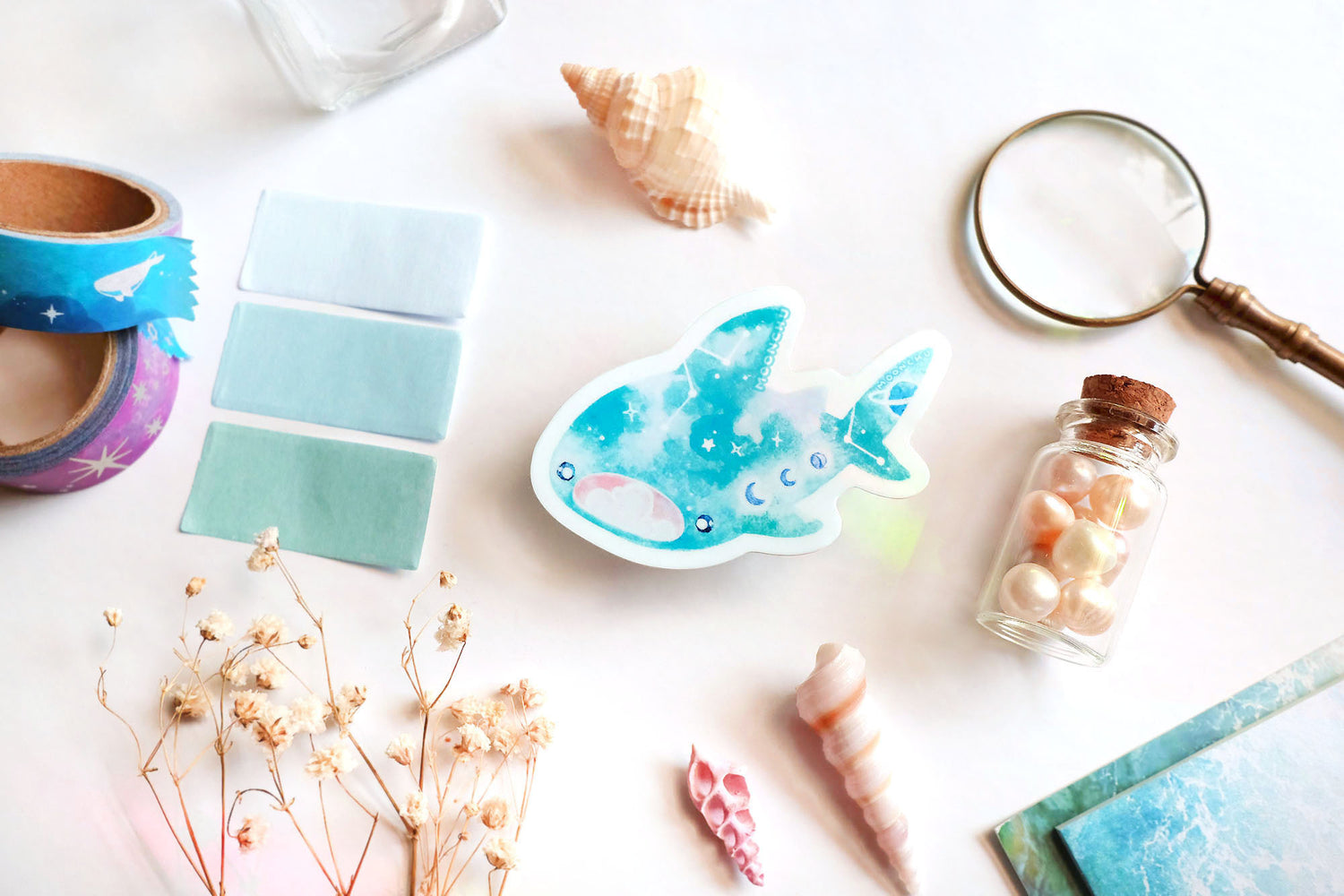 original art illustrated sticker by moonchu depicting a watercolour painted whale shark! mouth ajar, with colours of cyan and blue, it has little constellations and moon phases upon its body. it also has saturn on its tail! flatlay consists of other ocean items, like little shells and pearls. a high quality vinyl sticker for shark lovers or ocean gifts. perfect sticker for shark week.