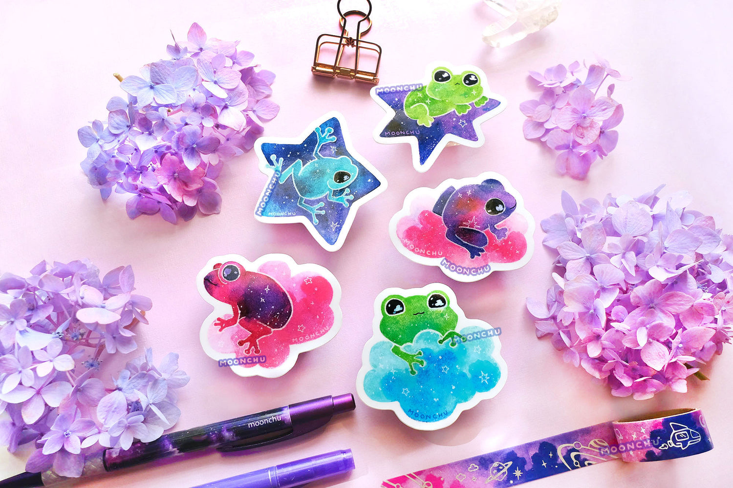 group photoshoot of vibrant original watercolour frog illustrations by moonchu, turned into glossy vinyl stickers. the cute frog friends are riding various purple and blue stars, pink clouds, and a big blue cloud. the amphibians range in colours from purples and pinks, to greens, and one teal. hydrangea blooms litter the backdrop. stickers would be perfect gifts for frog lovers to put on their water bottles, laptops, etc.
