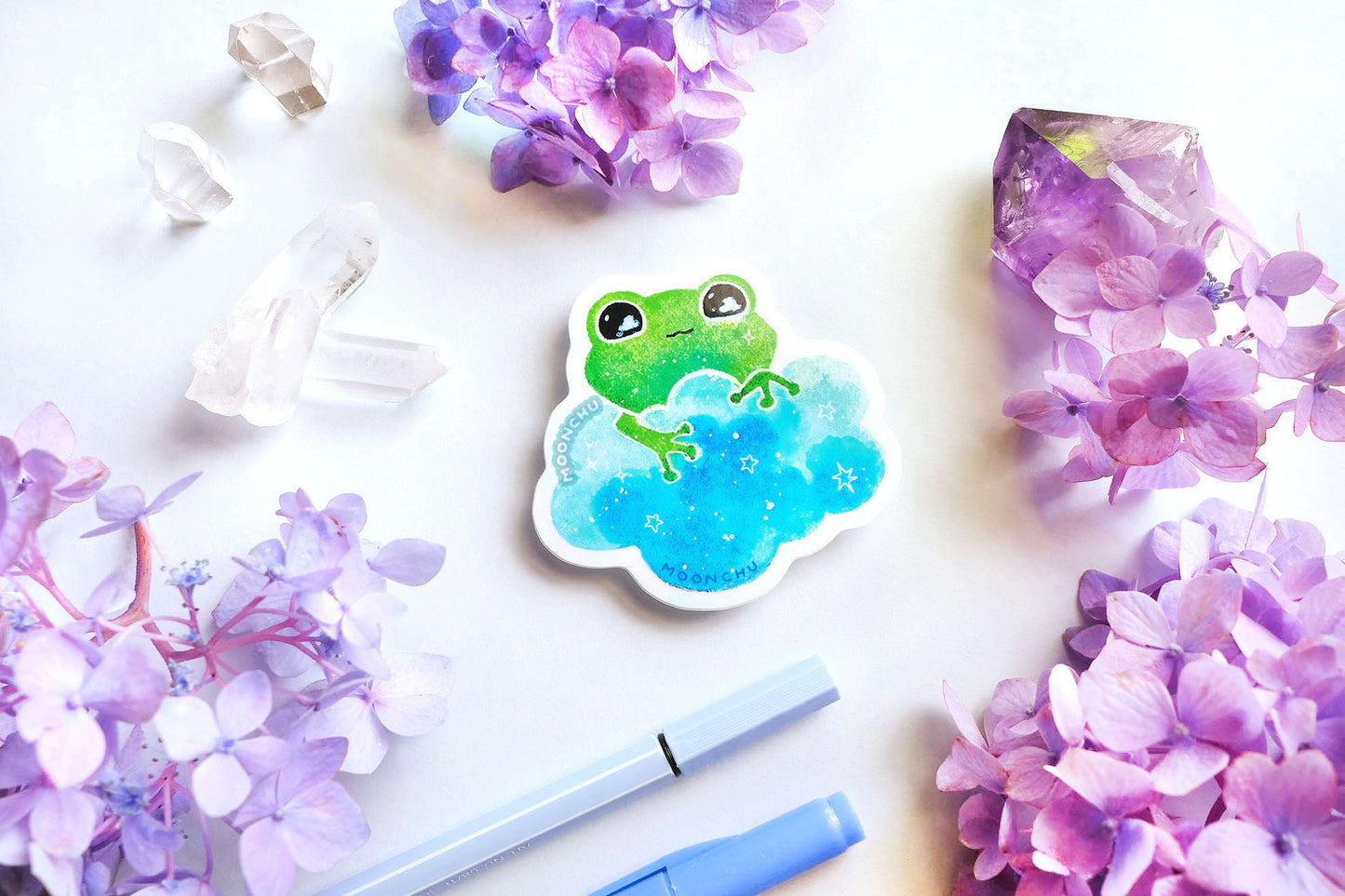 original watercolour painting by moonchu turned into a vinyl sticker! depicts a sweet green frog holding onto a sky blue cloud, with little stars twinkling and clouds in his eyes. quartz crystals and purple and blue hydrangea flowers litter the backdrop.