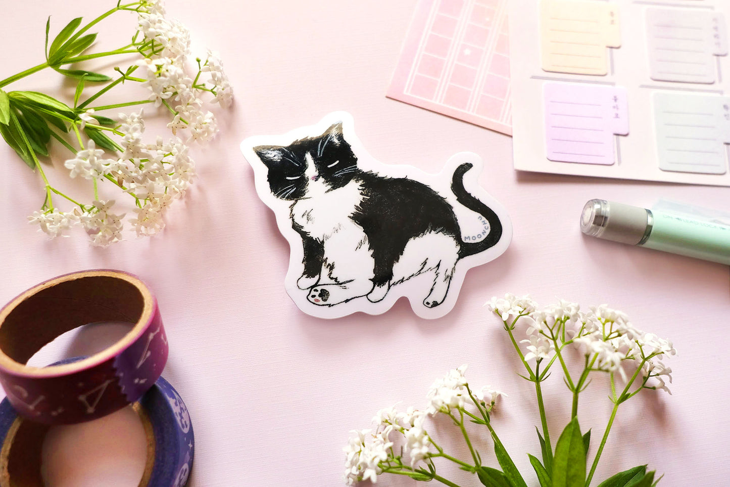 original art sticker by moonchu, illustrating a sweet little tuxedo cat. this high quality vinyl black kitty sticker is very glossy, and a tribute to mr. kitty. he is sitting with one of his signature legs stuck out. the rest of the flatlay consists of some stationery items and some flowers. a perfect sticker for tuxedo and black cat lovers!