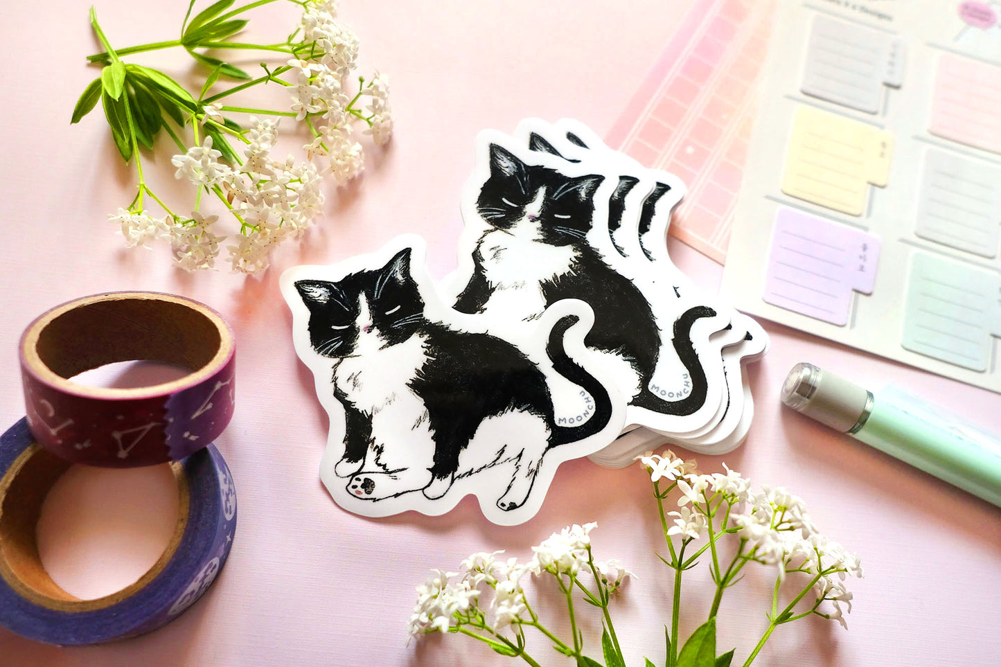 original art sticker by moonchu, illustrating a sweet little tuxedo cat. this high quality vinyl black kitty sticker is very glossy, and a tribute to mr. kitty. he is sitting with one of his signature legs stuck out. the rest of the flatlay consists of some stationery items and some flowers. a perfect sticker for tuxedo and black cat lovers!