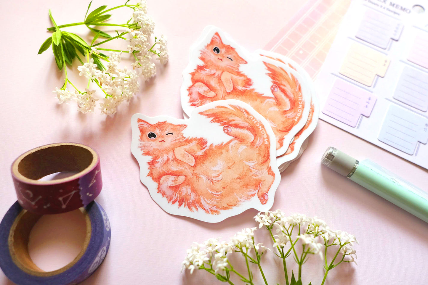original illustrated sticker by moonchu of her fluffy orange kitty! painted in watercolours, this high quality vinyl sticker illustrates a happy winky cat lying on his back. flatlay photo consists of pink stationery items and flowers. a sweet sticker for cat lovers and stationery lovers alike.