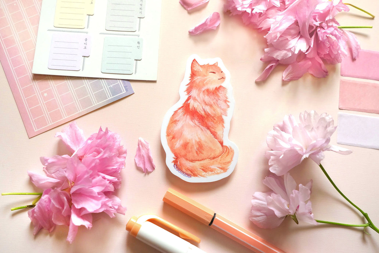 original illustrated sticker by moonchu of her fluffy orange kitty! painted in watercolours, this high quality vinyl sticker illustrates a regal, standing cat with his tail curled around his body. smiling, looking to his right. flatlay photo consists of pink stationery items and flowers. a sweet sticker for cat lovers and stationery lovers alike.