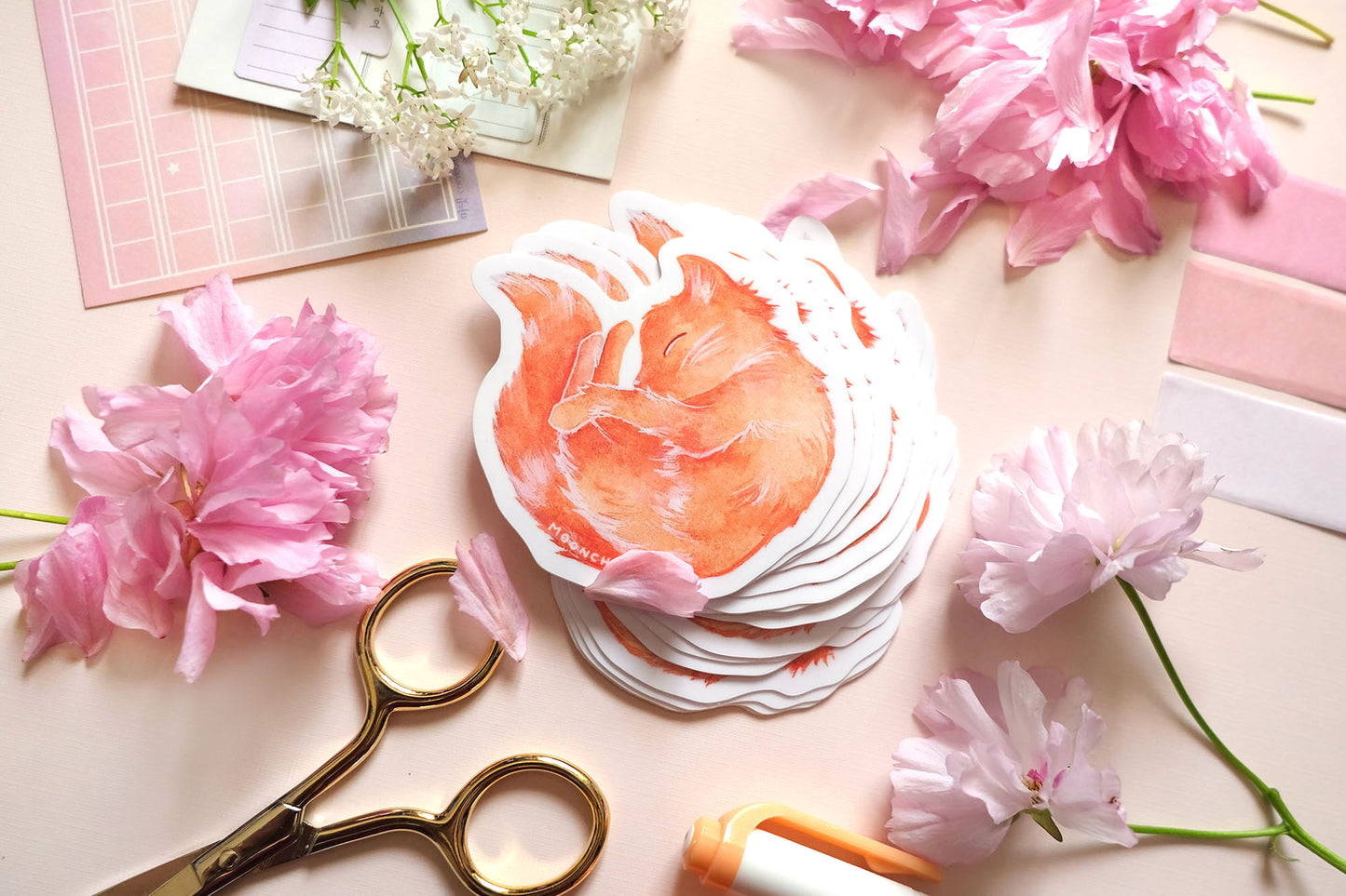 original sticker art by moonchu, watercolour orange cat painting, cat is curled up and sleepy like a donut. vibrant vinyl cat sticker is very cute, very orange! photographed on a flatlay consisting of pink stationery items and flowers.