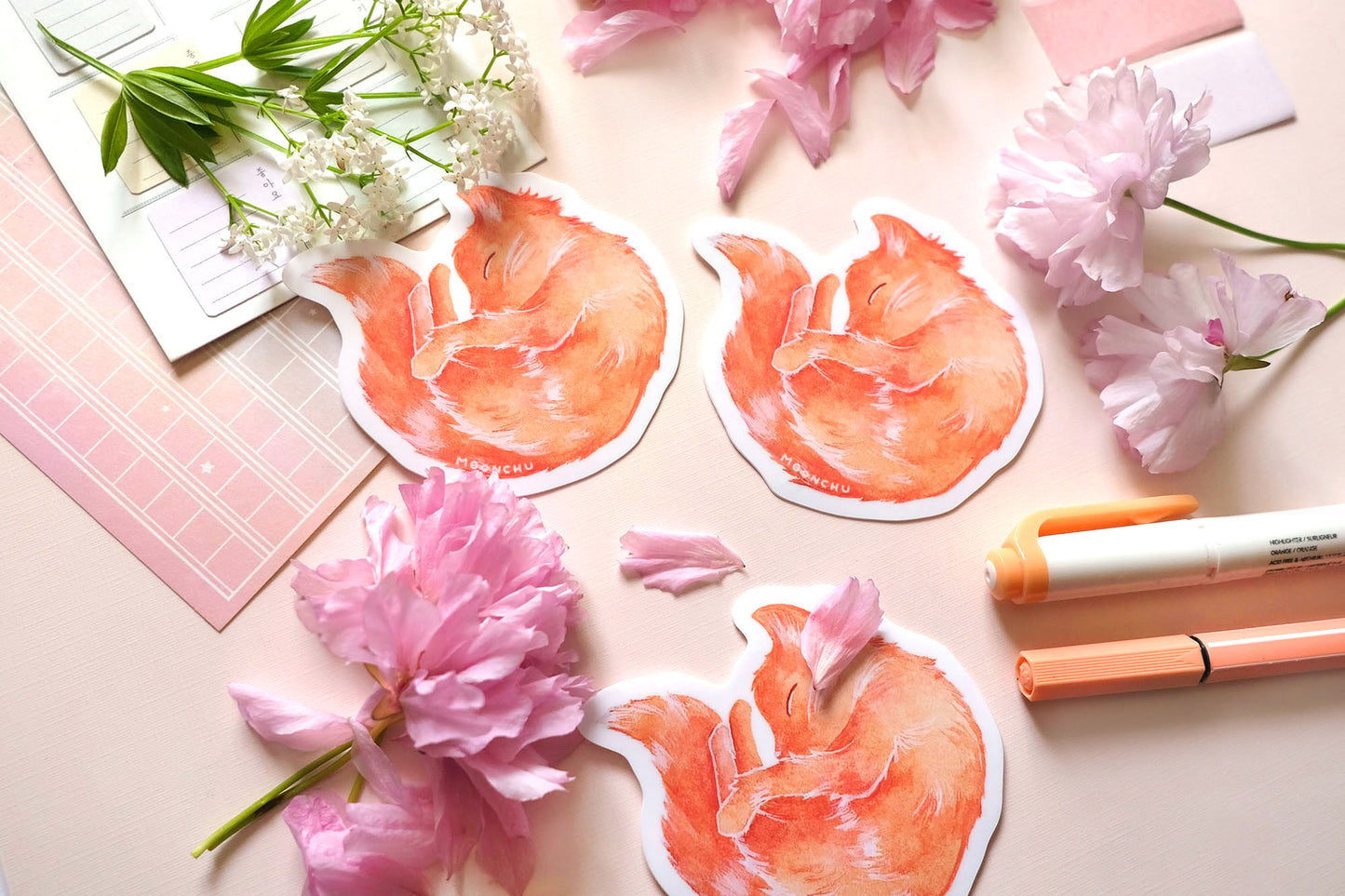 original sticker art by moonchu, watercolour orange cat painting, cat is curled up and sleepy like a donut. vibrant vinyl cat sticker is very cute, very orange! photographed on a flatlay consisting of pink stationery items and flowers.