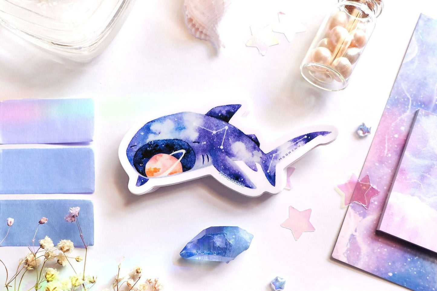original art illustrated sticker by moonchu depicting a watercolour painted megamouth shark! with a navy blue body, it has little constellations on its body. an orange saturn sits within its notorious open mouth. flatlay consists of other ocean items, like little shells and pearls. a high quality vinyl sticker for shark lovers or ocean gifts. perfect sticker for shark week.