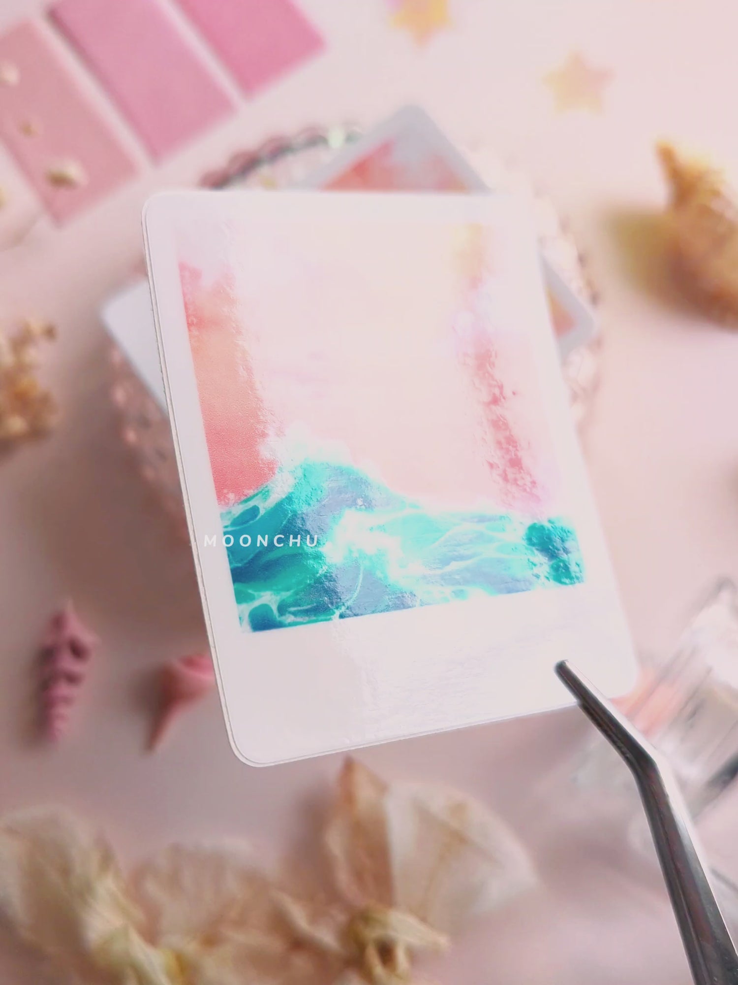 video showcasing moonchu's original art sticker depicting a beautiful orange sunset and a teal wave with a white border, like a printed snapshot. a super glossy and saturated high quality vinyl sticker of ocean wave sunset. various stationery items, dried florals, and seashells adorn the image. a unique sticker for any ocean lover or beach goer. would look great on a water bottle that you take to the beach during the summer!