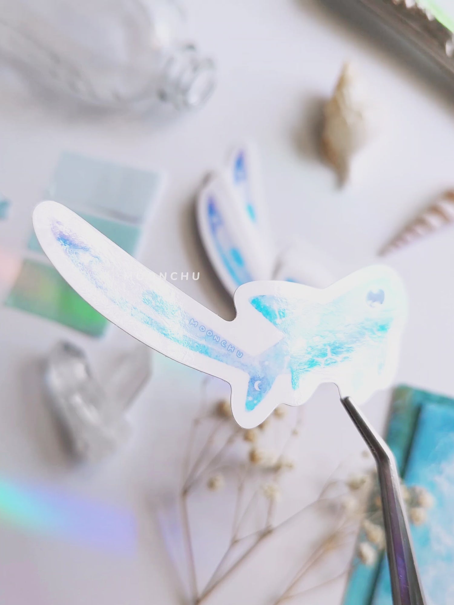 video showcases original art illustrated sticker by moonchu depicting a watercolour painted thresher shark! high gloss sticker. flatlay consists of other ocean items, like little shells and pearls. a high quality vinyl sticker for shark lovers or ocean gifts. perfect sticker for shark week.