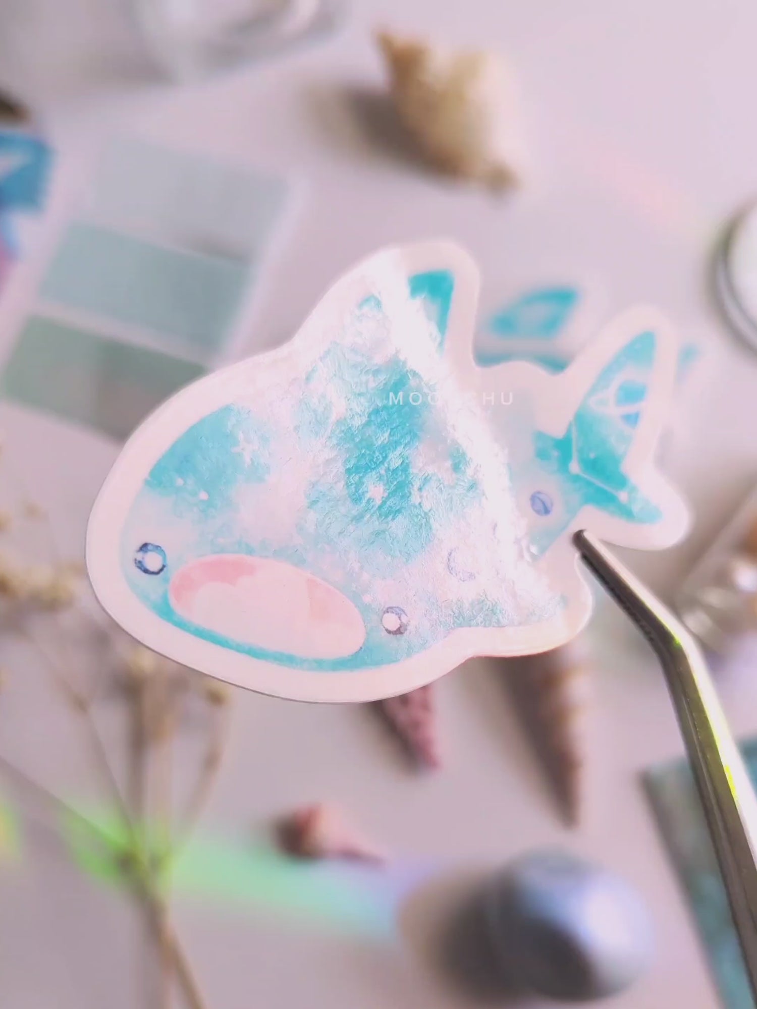 video showcases original art illustrated sticker by moonchu depicting a watercolour painted whale shark! mouth ajar, with colours of cyan and blue, it has little constellations and moon phases upon its body. it also has saturn on its tail! flatlay consists of other ocean items, like little shells and pearls. a high quality vinyl sticker for shark lovers or ocean gifts. perfect sticker for shark week.