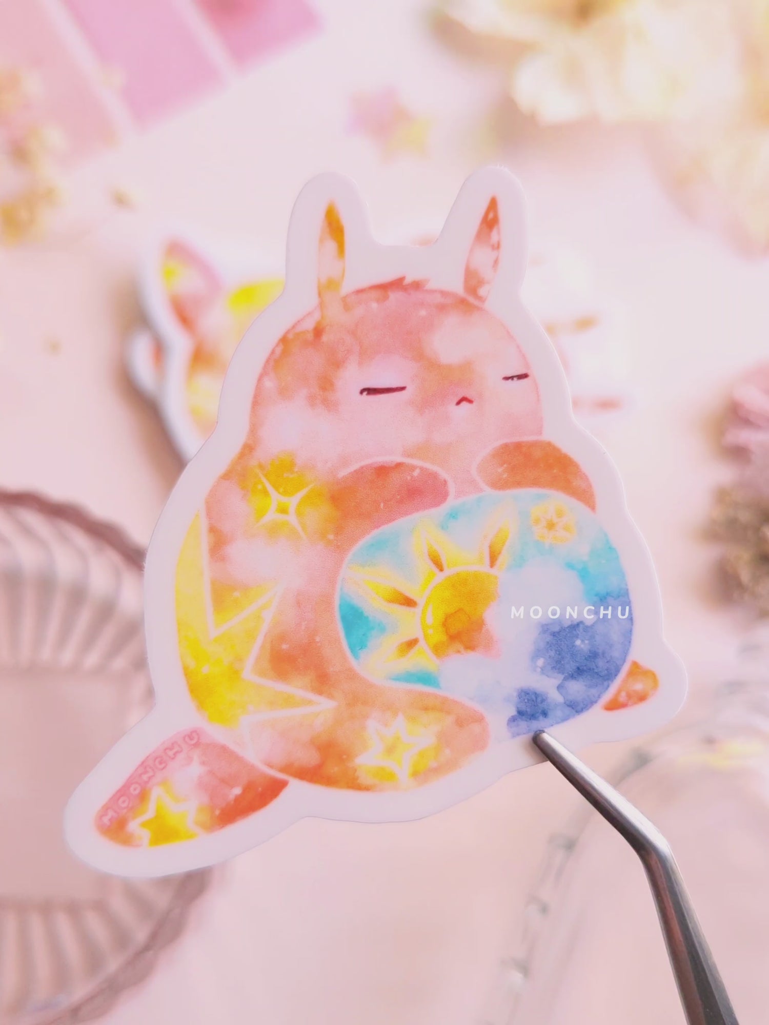 video is showcasing moonchu watercolor sticker depicting a v cute chubby bunny orange with yellow twinkling stars & a sun rising above blue clouds with a teal sky on his belly. he has sleepy and serene eyes. flowers, stars, and other items decorate the perimeter of sticker. a high quality colourful whimsical vinyl sticker. rest of the flatlay consists of other little pink stationery items, stars, and dried flowers.