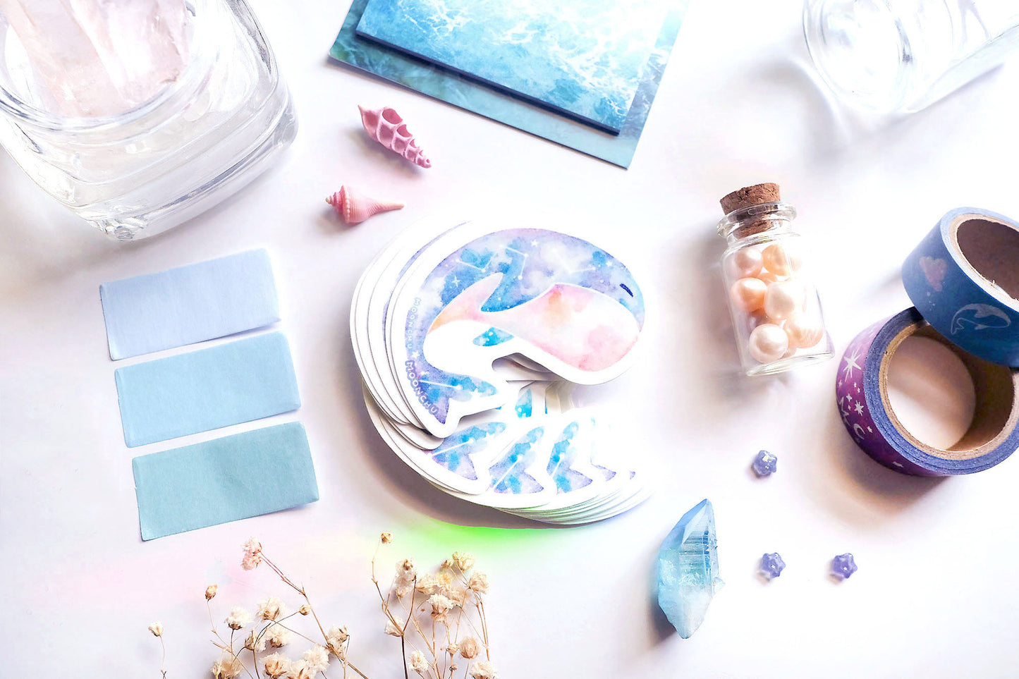 original art illustrated sticker by moonchu depicting a watercolour painted calming whale! with a serene cyan, blue, and pink gradient body and little constellations. has a sleepy face and a pink upturned belly. flatlay consists of other ocean items, like little shells and pearls. a high quality vinyl sticker for shark lovers or ocean gifts. perfect sticker for shark week.