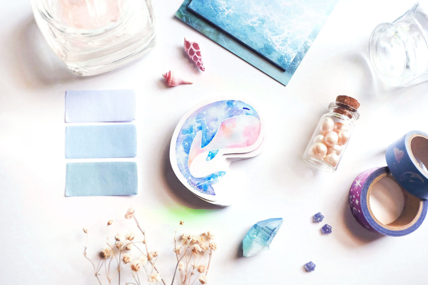 original art illustrated sticker by moonchu depicting a watercolour painted calming whale! with a serene cyan, blue, and pink gradient body and little constellations. has a sleepy face and a pink upturned belly. flatlay consists of other ocean items, like little shells and pearls. a high quality vinyl sticker for shark lovers or ocean gifts. perfect sticker for shark week.