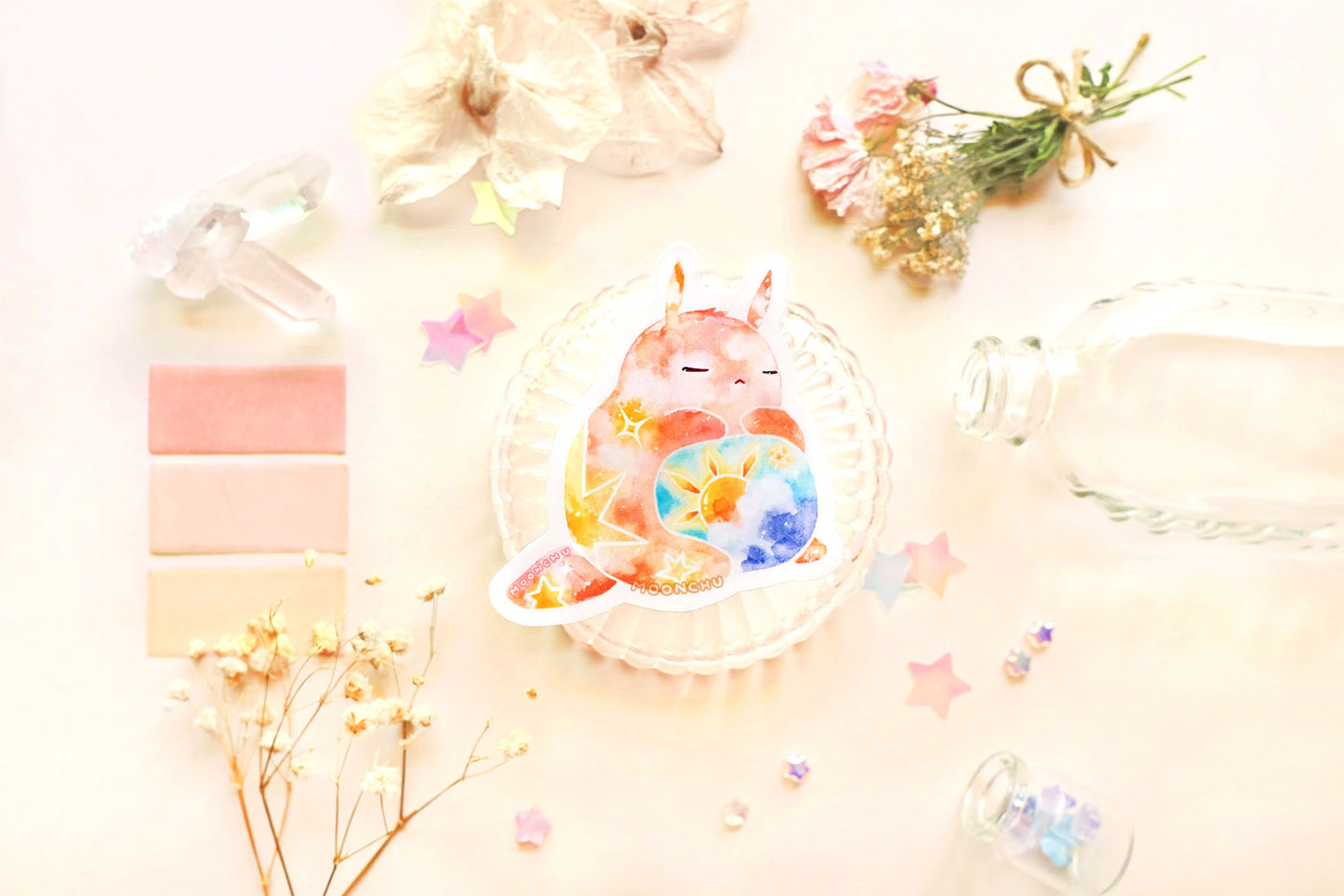 photo is of moonchu watercolor sticker depicting a v cute chubby bunny orange with yellow twinkling stars & a sun rising above blue clouds with a teal sky on his belly. he has sleepy and serene eyes. flowers, stars, and other items decorate the perimeter of sticker. a high quality colourful whimsical vinyl sticker. rest of the flatlay consists of other little pink stationery items, stars, and dried flowers.
