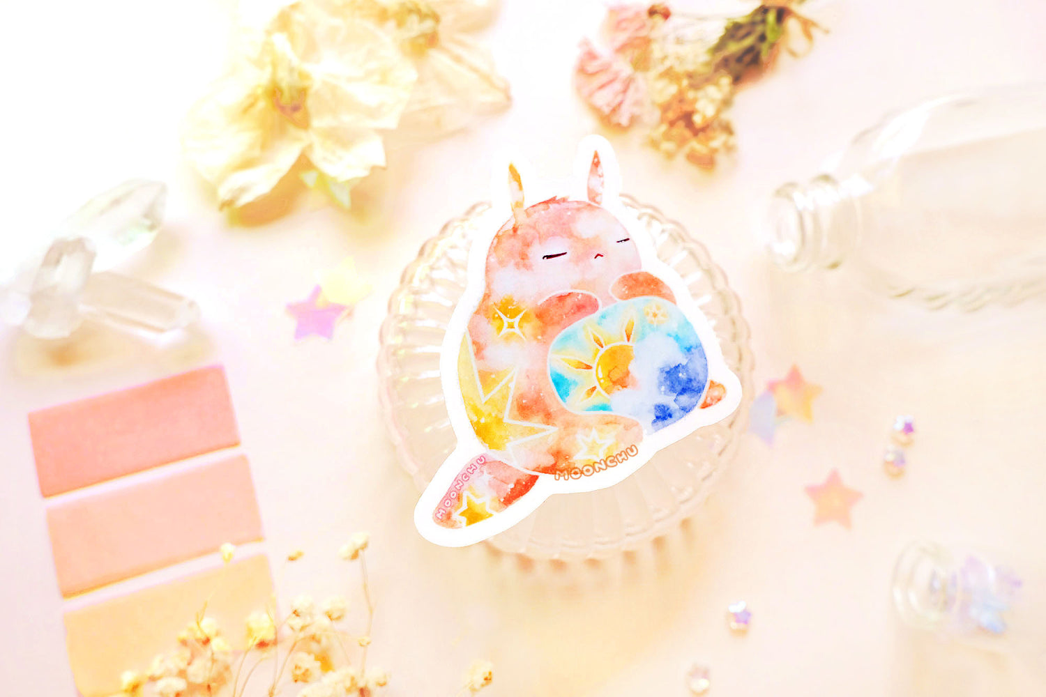 photo is of moonchu watercolor sticker depicting a v cute chubby bunny orange with yellow twinkling stars & a sun rising above blue clouds with a teal sky on his belly. he has sleepy and serene eyes. flowers, stars, and other items decorate the perimeter of sticker. a high quality colourful whimsical vinyl sticker. rest of the flatlay consists of other little pink stationery items, stars, and dried flowers.