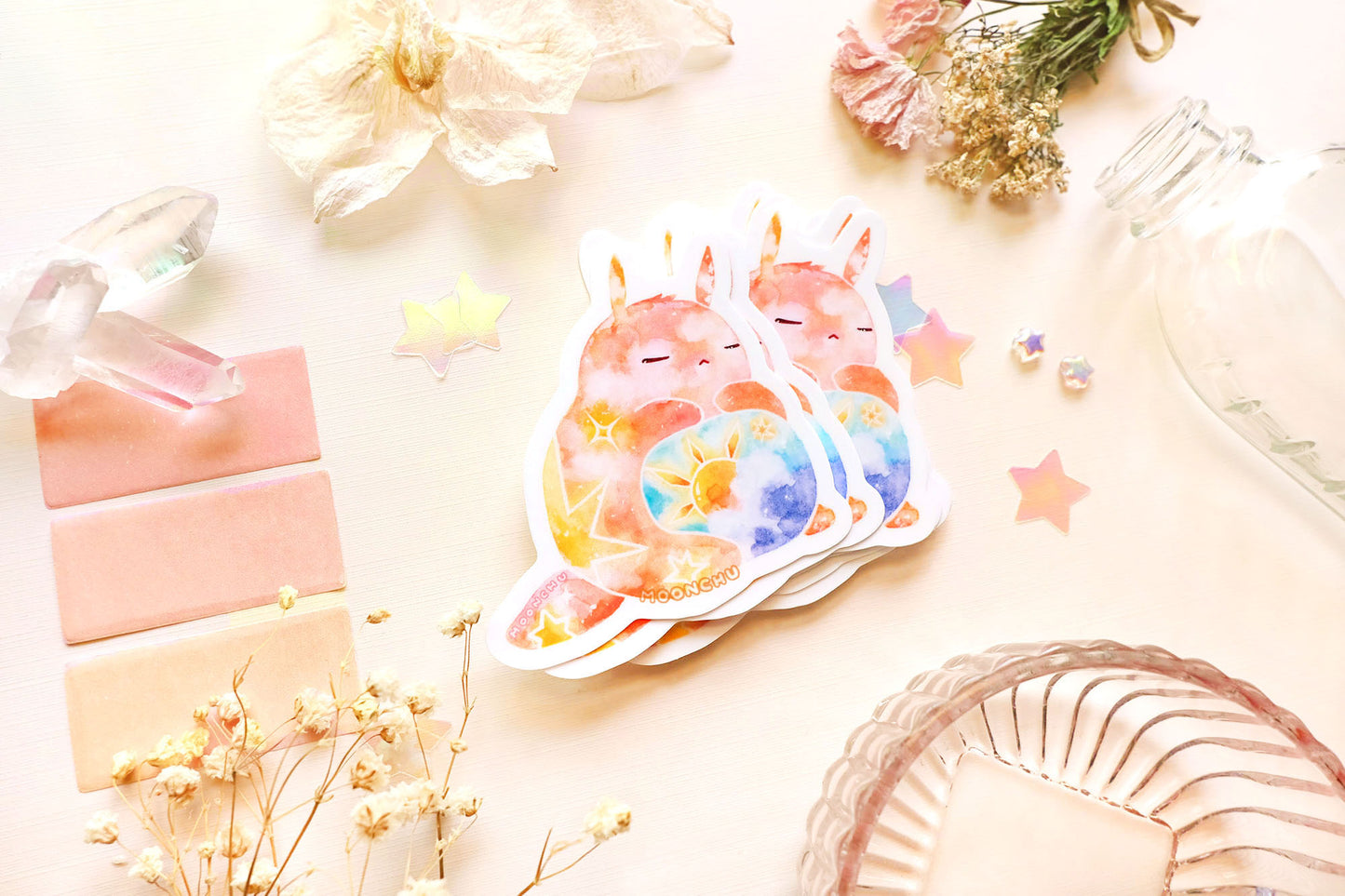 photo is of moonchu watercolor sticker depicting a v cute chubby bunny orange with yellow twinkling stars & a sun rising above blue clouds with a teal sky on his belly. he has sleepy and serene eyes. flowers, stars, and other items decorate the perimeter of sticker. a high quality colourful whimsical vinyl sticker. rest of the flatlay consists of other little pink stationery items, stars, and dried flowers.