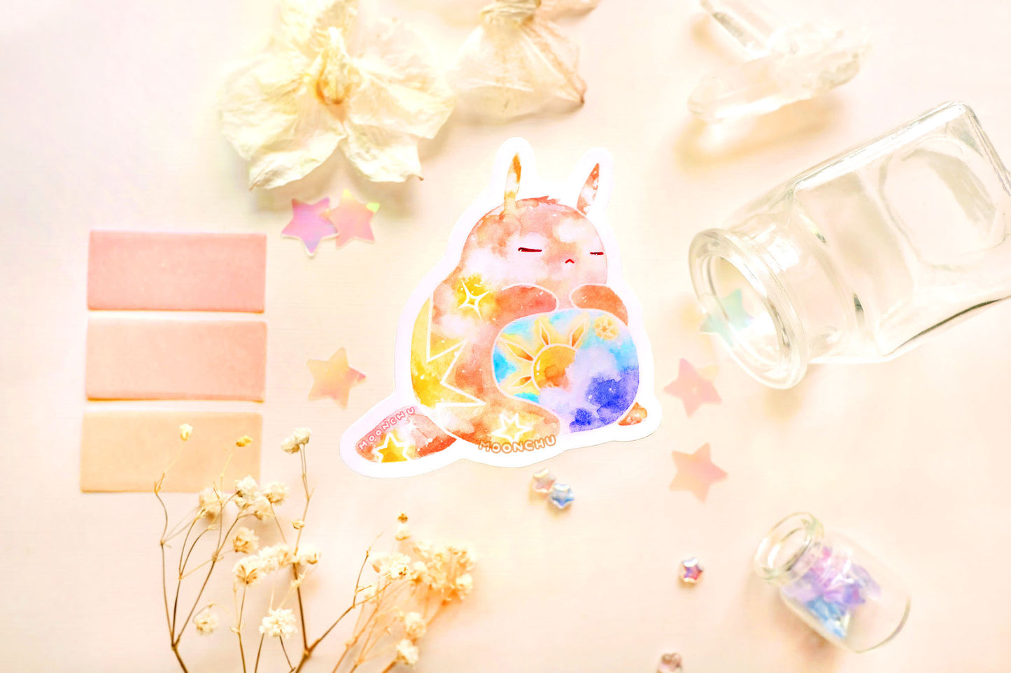 photo is of moonchu watercolor sticker depicting a v cute chubby bunny orange with yellow twinkling stars & a sun rising above blue clouds with a teal sky on his belly. he has sleepy and serene eyes. flowers, stars, and other items decorate the perimeter of sticker. a high quality colourful whimsical vinyl sticker. rest of the flatlay consists of other little pink stationery items, stars, and dried flowers.