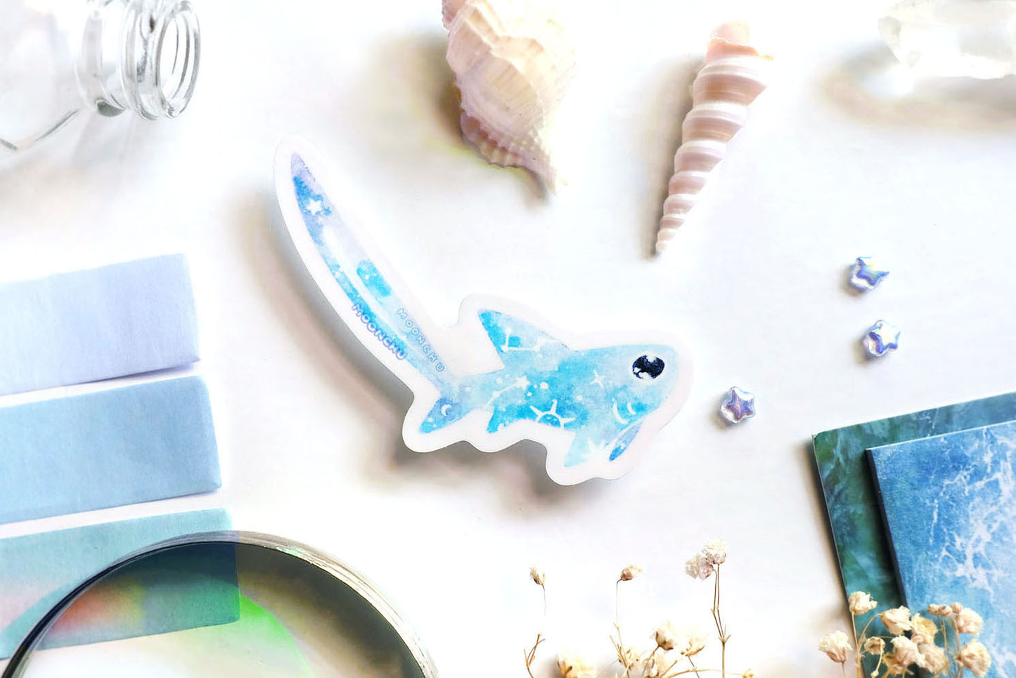 original art illustrated sticker by moonchu depicting a watercolour painted thresher shark! with a light blue body, it has little constellations on its body and a shooting star on its notoriously large tail. flatlay consists of other ocean items, like little shells and pearls. a high quality vinyl sticker for shark lovers or ocean gifts. perfect sticker for shark week.