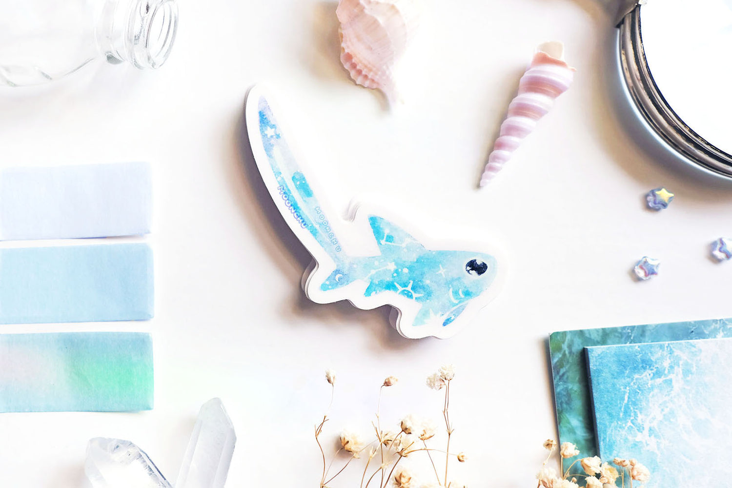 original art illustrated sticker by moonchu depicting a watercolour painted thresher shark! with a light blue body, it has little constellations on its body and a shooting star on its notoriously large tail. flatlay consists of other ocean items, like little shells and pearls. a high quality vinyl sticker for shark lovers or ocean gifts. perfect sticker for shark week.