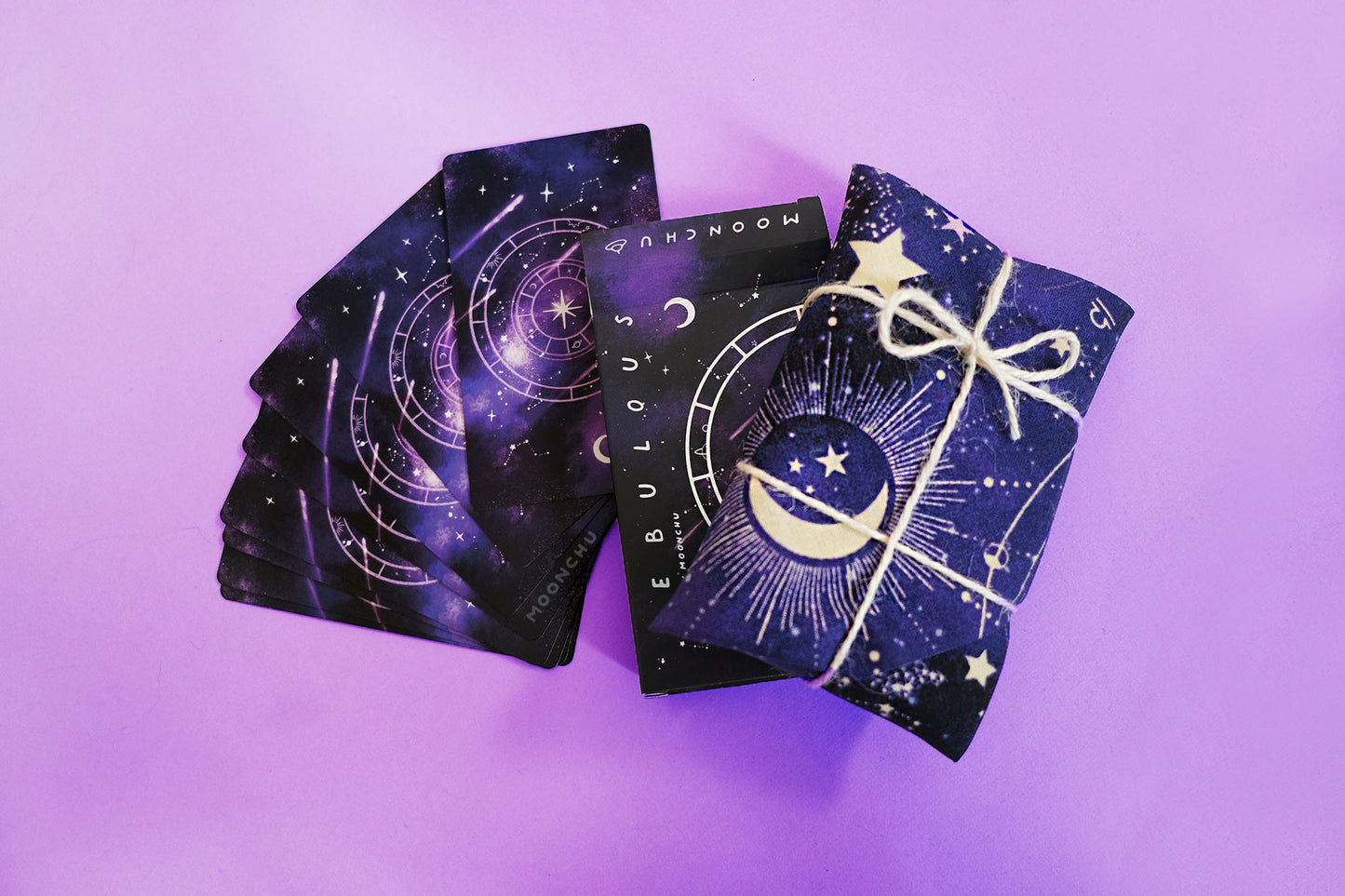 stunning indie holographic tarot deck by moonchu with all original art designs! this photograph depicts the holographic detailed tarot box design, with a galactic theme of a rich purple background and shooting stars originally painted in watercolour. this is accented by graphic designs of a crescent moon, nebulous tarot major arcana name, and intricate wheel design - all in holographic foil. comes lovingly hand-wrapped in a blue galaxy space themed cloth and twine string.