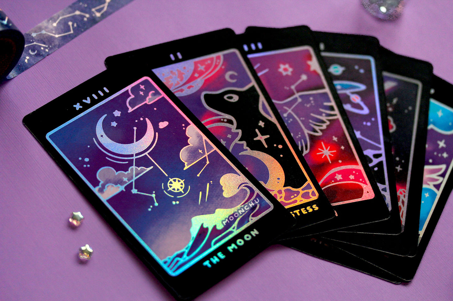 holo indie major arcana tarot deck by moonchu, all original art designs! featuring several magical nature and animal themed tarot cards. photo depicts the moon, a popular moonchu original tarot design with a crescent moon hung amongst clouds, constellations, and above an ocean wave. all with strategic delicate holographic accents. other tarot card previews of the high priestess (wolves), death (crow), the hanged man (whale shark), the tower (crystal), and temperance (winged cat).