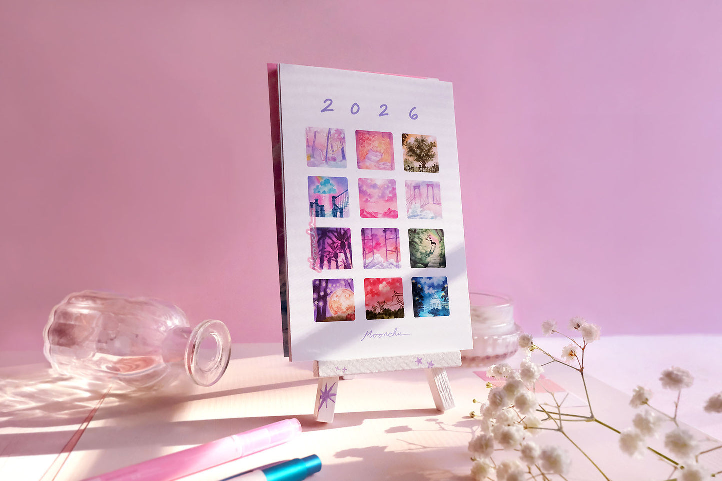 showcases previews of the 12 monthly original art paintings that moonchu did for the calendar. each one is a small square with rounded edges. 2026 text at top and "moonchu" script at bottom. preview images are a rainbow of colour. loose-leaf calendar is perched upon a white, hand-painted easel with purple stars. various objects line the rest of the photo for a pretty atmosphere.