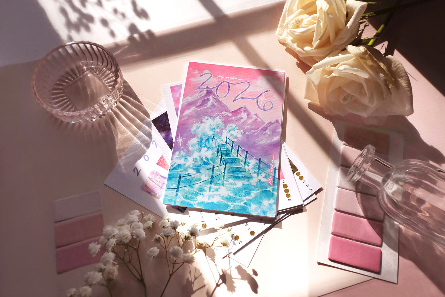 through a sunstreamed window, there's a flatlay of flowers, stationery items, glass, and moonchu's original art 2026 calendar. showcases the cover art which has a custom 2026 cursive script, a pinky peach sunset with some clouds overlooking a purple mountain peak, and a foamy sea wave of teal splashing below onto a teal set of stairs. can see hand-embellished golden moon phase stickers on other months.