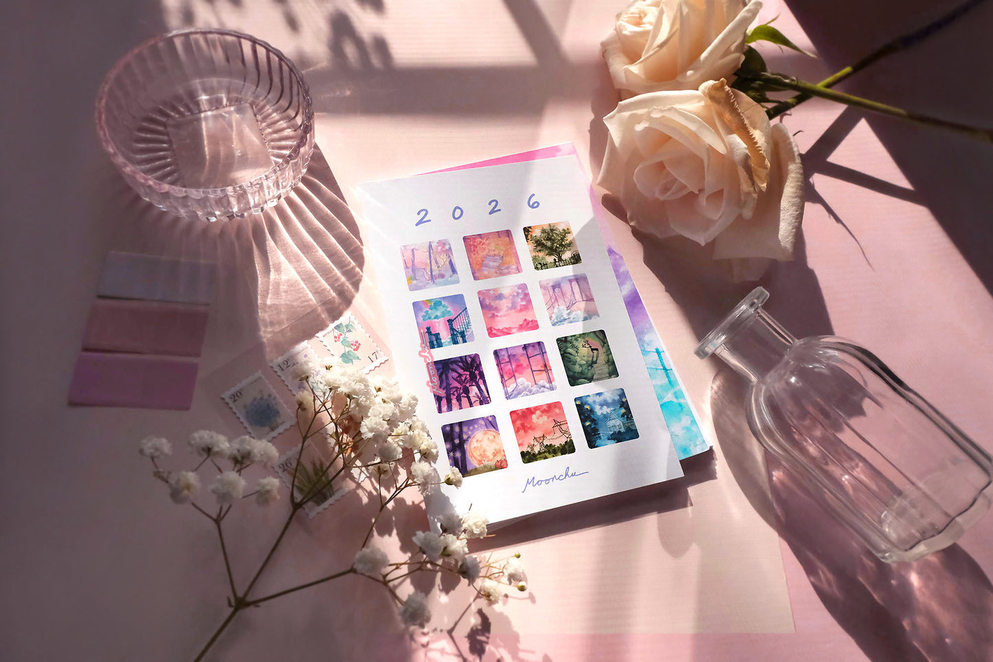 showcases previews of the 12 monthly original art paintings that moonchu did for the calendar. each one is a small square with rounded edges. 2026 text at top and "moonchu" script at bottom. preview images are a rainbow of colour. loose-leaf calendar is perched upon a white, hand-painted easel with purple stars. various objects line the rest of the photo for a pretty atmosphere.