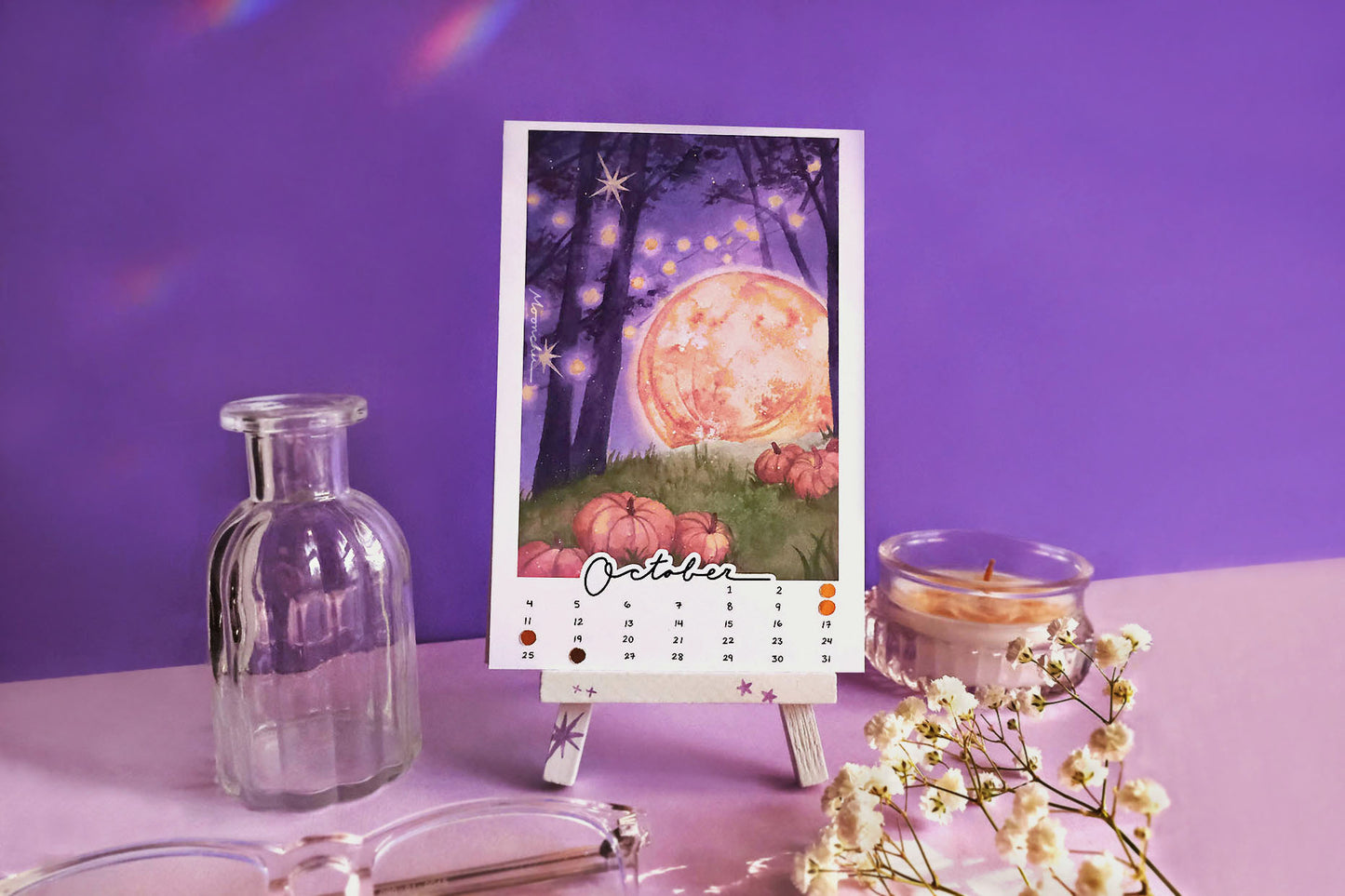 the month of october for the moonchu calendar. this original watercolour painting is comprised of a dark purple forest with yellow fairy lights draped between the trees. a large, yellow full moon glows in the right side of the painting. a lush grassy plain holds several orange pumpkins. inspired by the great pumpkin! october is in cursive script at the bottom, hand-written numbers date the month. hand-placed golden circles notate the moon phases of the month.