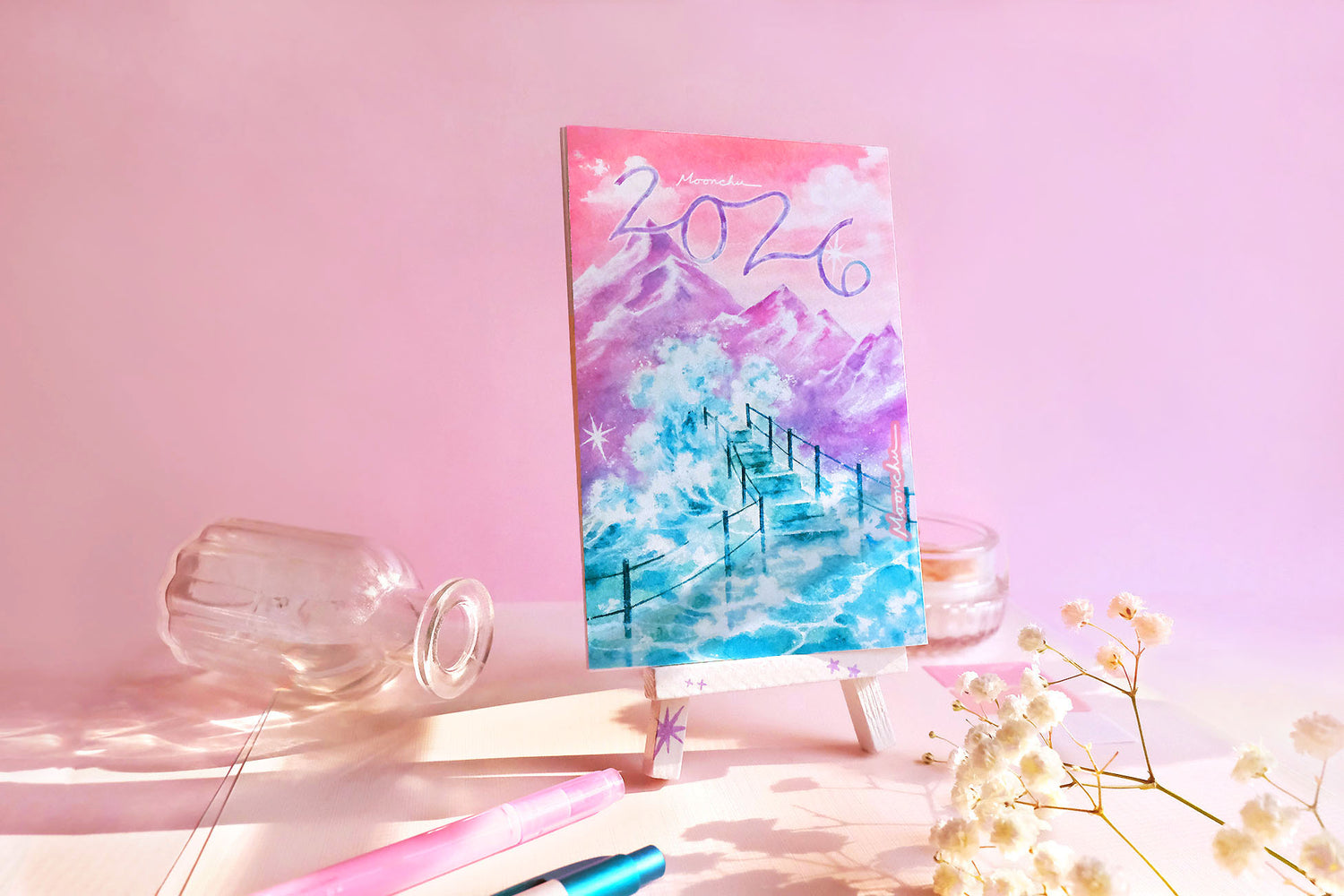 showcases the cover art of moonchu's 2026 original art calendar which has a custom 2026 cursive script, a pinky peach sunset with some clouds overlooking a purple mountain peak, and a foamy sea wave of teal splashing below onto a teal set of stairs. loose leaf calendar perches upon a hand-painted white easel with purple stars, amongst other fun objects like glass items, a pink and blue pen, and some flowers.
