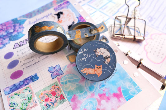 original artist washi tape by moonchu as a tribute to two of her cats, akki and maple. this photo showcases the packaging label with one illustration of akki and one illustration of maple on a navy/blue background with white cloud and star doodles. the rest of the flatlay consists of other cute stationery items and journal pages.