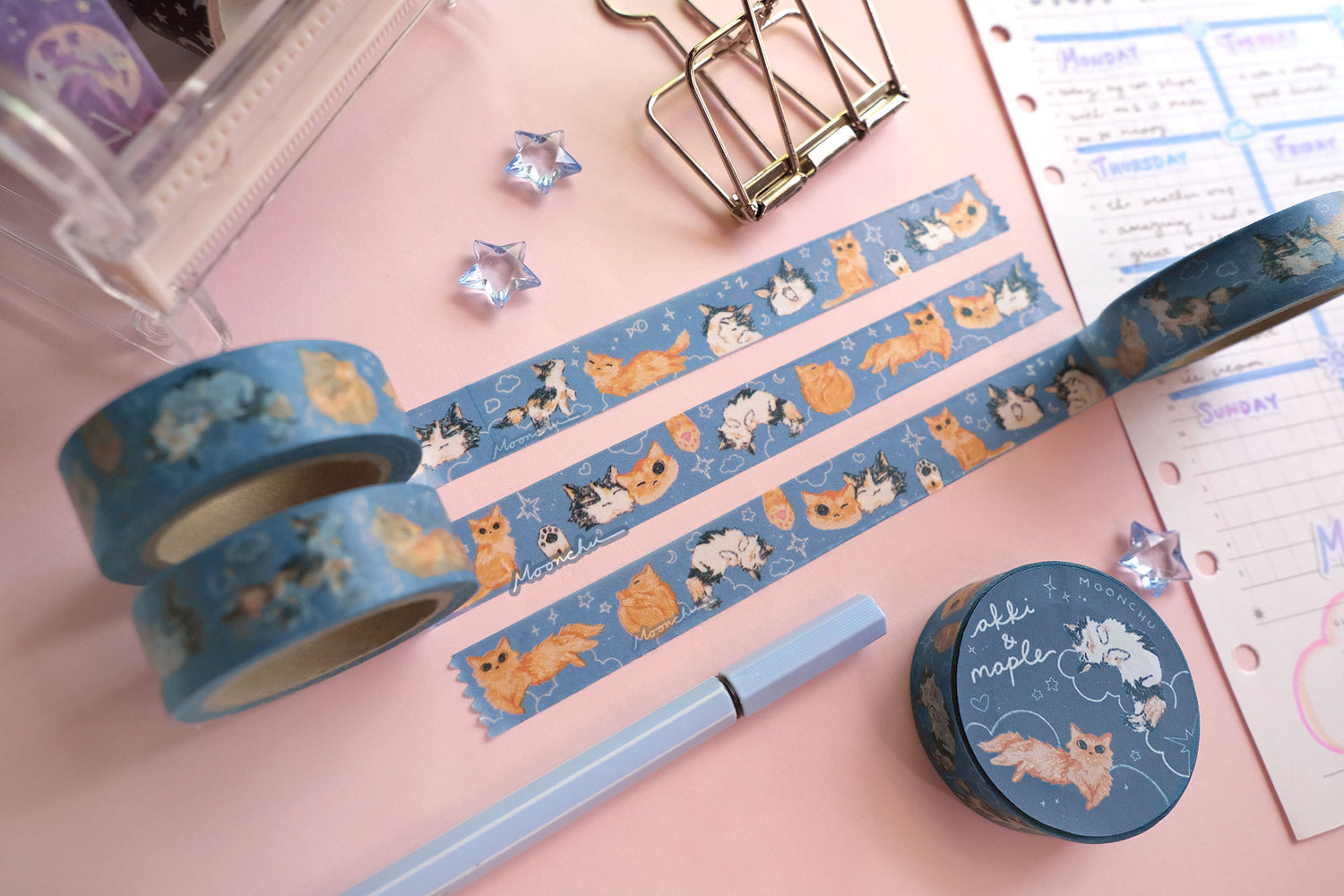 original artist washi tape by moonchu as a tribute to two of her cats, akki and maple. three washi tape are unrolled for the viewer to see the various kitty designs (both of their faces smushed together and smiling, a calico paw and an orange kitty paw, akki and maple in sleeping/sitting/yawning poses). orange cat and calico cat are drawn in various poses on a navy/blue background with white doodles (such as clouds and stars and hearts). the rest of the flatlay consists of other cute stationery items.