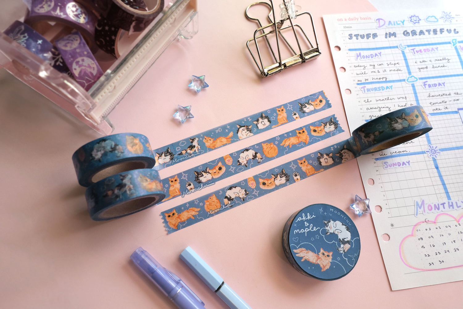 original artist washi tape by moonchu as a tribute to two of her cats, akki and maple. three washi tape are unrolled for the viewer to see the various kitty designs (both of their faces smushed together and smiling, a calico paw and an orange kitty paw, akki and maple in sleeping/sitting/yawning poses). orange cat and calico cat are drawn on a navy/blue background with white doodles (such as clouds and stars and hearts). the rest of the flatlay consists of other cute stationery items.