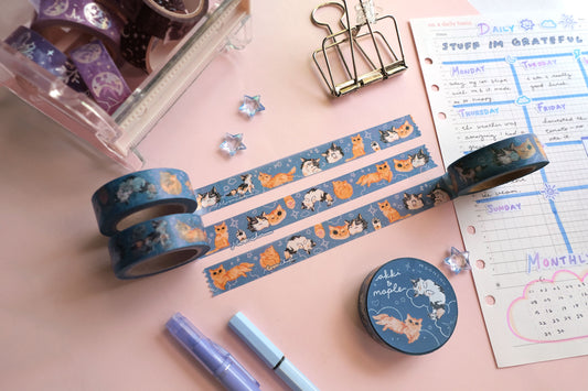 original artist washi tape by moonchu as a tribute to two of her cats, akki and maple. three washi tape are unrolled for the viewer to see the various kitty designs (both of their faces smushed together and smiling, a calico paw and an orange kitty paw, akki and maple in sleeping/sitting/yawning poses). orange cat and calico cat are drawn on a navy/blue background with white doodles (such as clouds and stars and hearts). the rest of the flatlay consists of other cute stationery items.
