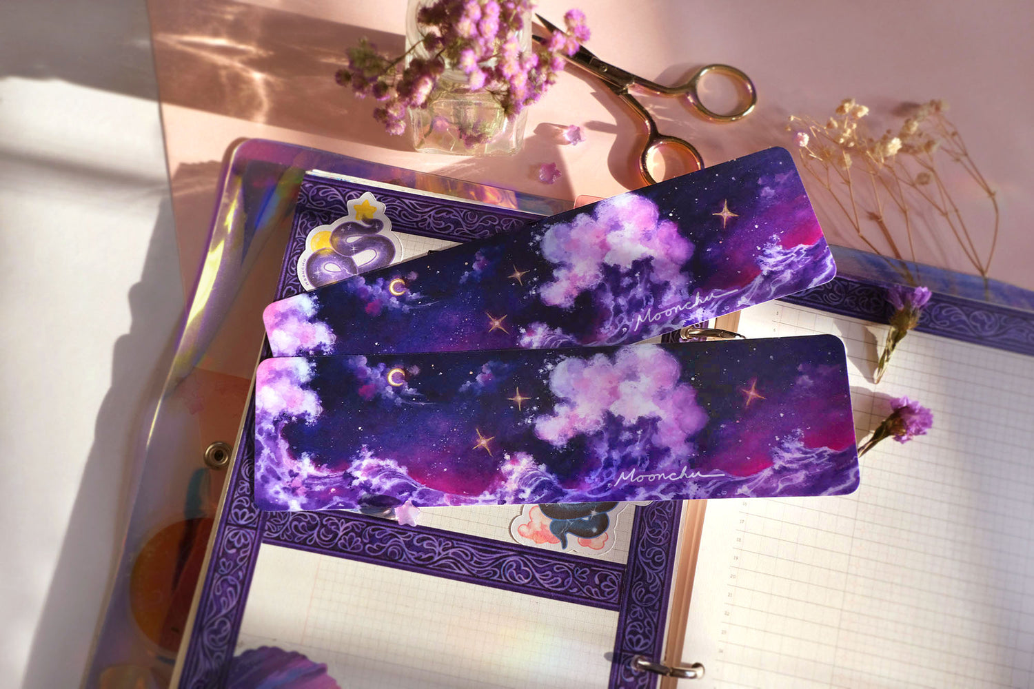 photo depicts flatlay image with moonchu original art bookmark illustrating a rich blue, purple, and magenta night sky with misty clouds and crescent moon. three twinkling stars are hanging in the sky, which are accented with metallic gold. beneath the sky lies a fluffy amethyst wave, crashing against each other. a beautifully saturated full colour bookmark. glossy laminated bookmark for a bookish lover. original art bookmark lies upon an open novel, with purple dried flora & purple pens.