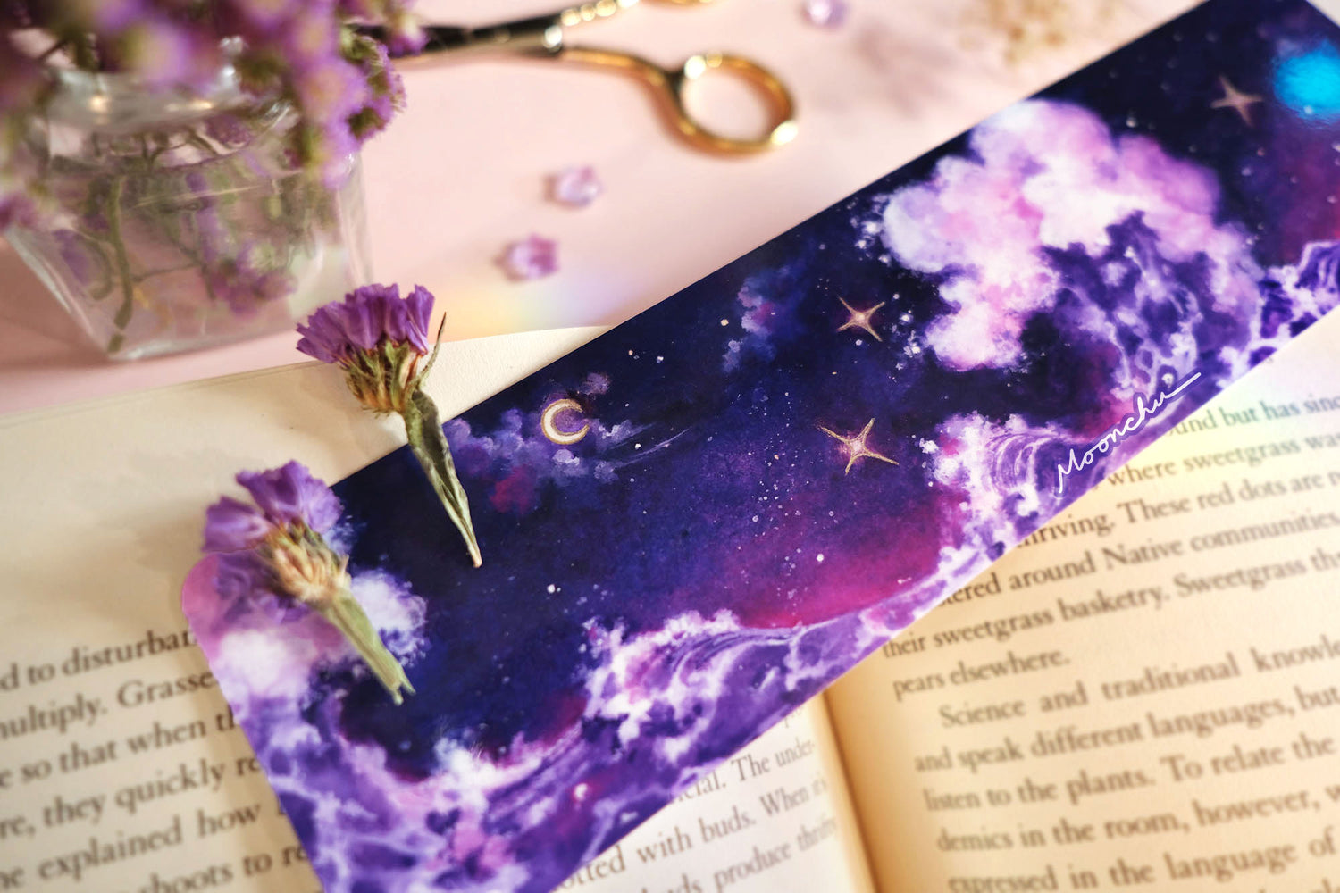 photo depicts flatlay image with moonchu original art bookmark illustrating a rich blue, purple, and magenta night sky with misty clouds and crescent moon. three twinkling stars are hanging in the sky, which are accented with metallic gold. beneath the sky lies a fluffy amethyst wave, crashing against each other. a beautifully saturated full colour bookmark. glossy laminated bookmark for a bookish lover. original art bookmark lies upon an open novel, with purple dried flora & purple pens.