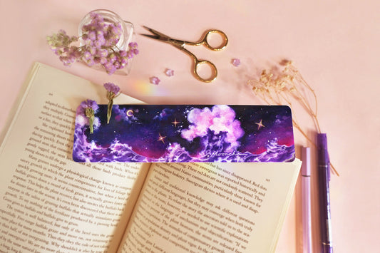 photo depicts flatlay image with moonchu original art bookmark illustrating a rich blue, purple, and magenta night sky with misty clouds and crescent moon. three twinkling stars are hanging in the sky, which are accented with metallic gold. beneath the sky lies a fluffy amethyst wave, crashing against each other. a beautifully saturated full colour bookmark. glossy laminated bookmark for a bookish lover. original art bookmark lies upon an open novel, with purple dried flora & purple pens.