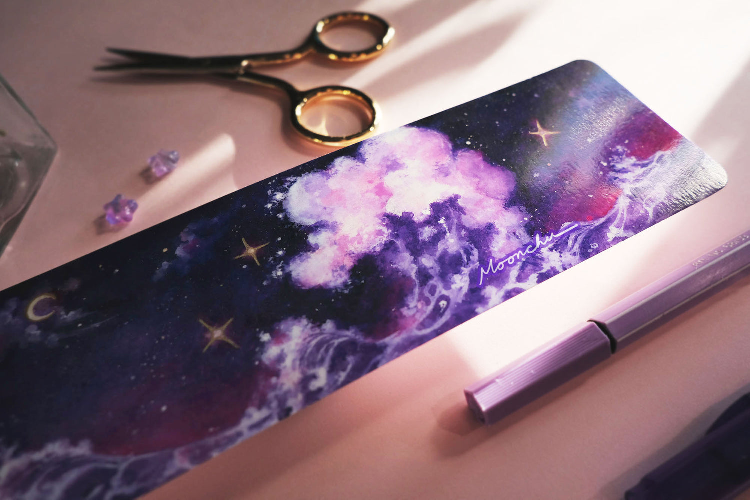photo depicts flatlay image with moonchu original art bookmark illustrating a rich blue, purple, and magenta night sky with misty clouds and crescent moon. three twinkling stars are hanging in the sky, which are accented with metallic gold. beneath the sky lies a fluffy amethyst wave, crashing against each other. a beautifully saturated full colour bookmark. glossy laminated bookmark for a bookish lover. original art bookmark lies upon an open novel, with purple dried flora & purple pens.