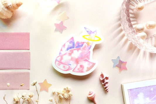 original art illustrated sticker by moonchu depicting a watercolour painted pastel angelshark! pretty pink, purple, and white clouds make up its body, as it smiles and inhales more clouds. a yellow halo rings around its tail. flatlay consists of other ocean items, like little shells and pearls. a high quality vinyl sticker for shark lovers or ocean gifts. perfect sticker for shark week.