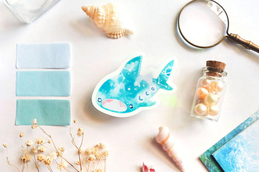 original art illustrated sticker by moonchu depicting a watercolour painted whale shark! mouth ajar, with colours of cyan and blue, it has little constellations and moon phases upon its body. it also has saturn on its tail! flatlay consists of other ocean items, like little shells and pearls. a high quality vinyl sticker for shark lovers or ocean gifts. perfect sticker for shark week.