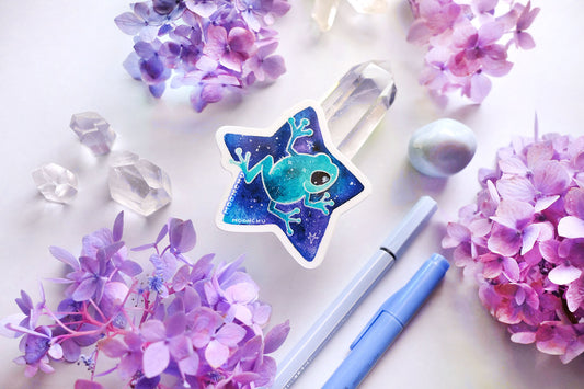 original watercolour painting by moonchu turned into a vinyl sticker! depicts a teal frog riding a saturated blue cosmic star. quartz crystals and purple and blue hydrangea flowers litter the backdrop. a cool sticker for frog enthusiasts and space lovers.