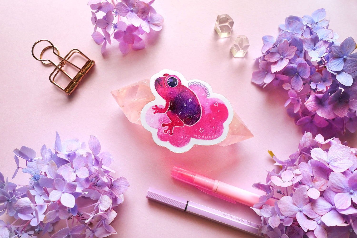 original watercolour painting by moonchu turned into a vinyl sticker! depicts a jammy strawberry magenta and purple frog, smiling and sitting on a puffy pink cloud. he is looking to the left, maybe thinking about a snack. little stars are scattered amongst the sticker. quartz crystals and purple and blue hydrangea flowers litter a pink backdrop.