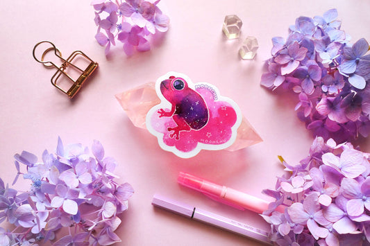 original watercolour painting by moonchu turned into a vinyl sticker! depicts a jammy strawberry magenta and purple frog, smiling and sitting on a puffy pink cloud. he is looking to the left, maybe thinking about a snack. little stars are scattered amongst the sticker. quartz crystals and purple and blue hydrangea flowers litter a pink backdrop. great sticker for frog lovers.