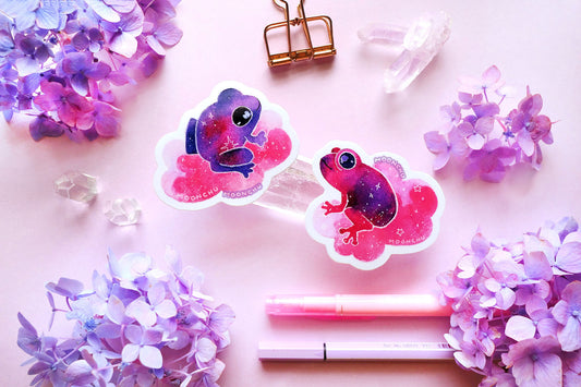 original watercolour painting by moonchu turned into vibrant vinyl stickers! depicts two little froggy friend stickers. one is purple with some pink, and the other is pink with some purple, and both are riding puffy pink clouds. they are facing each other, probably bonded soul frogs. little stars are scattered amongst the stickers. quartz crystals and purple and blue hydrangea flowers litter a pink backdrop.