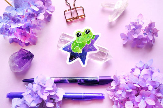 original watercolour painting by moonchu turned into a vinyl sticker! depicts a sweet green frog perched atop a purple star, blasting off on an adventure! little twinkling stars are on its body, as well as the star itself. quartz crystals and purple and blue hydrangea flowers litter the backdrop.
